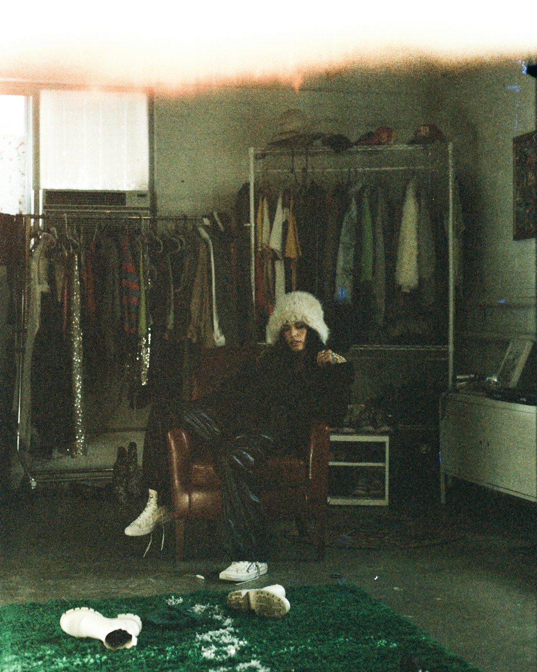 Person in fluffy hat sits in armchair surrounded by clothes.